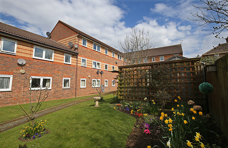 Landscaped communal garden beside a three‑storey red‑brick residential building with white‑framed windows. A well‑kept lawn runs through the centre, bordered by flower beds filled with daffodils and colourful planting, with a wooden trellis fence and shrubs to one side under a partly cloudy blue sky.