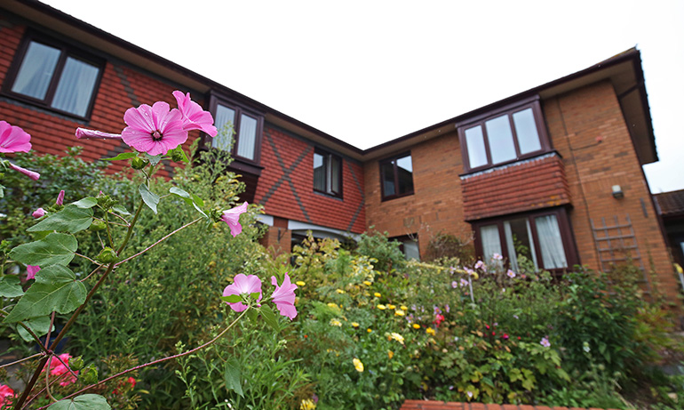 Exterior of a two‑storey red‑brick residential building with wooden‑framed windows, viewed over a densely planted garden in the foreground. Pink flowers and green foliage fill raised beds closest to the camera, with the building’s upper windows and timber‑style detailing visible behind the planting under an overcast sky.