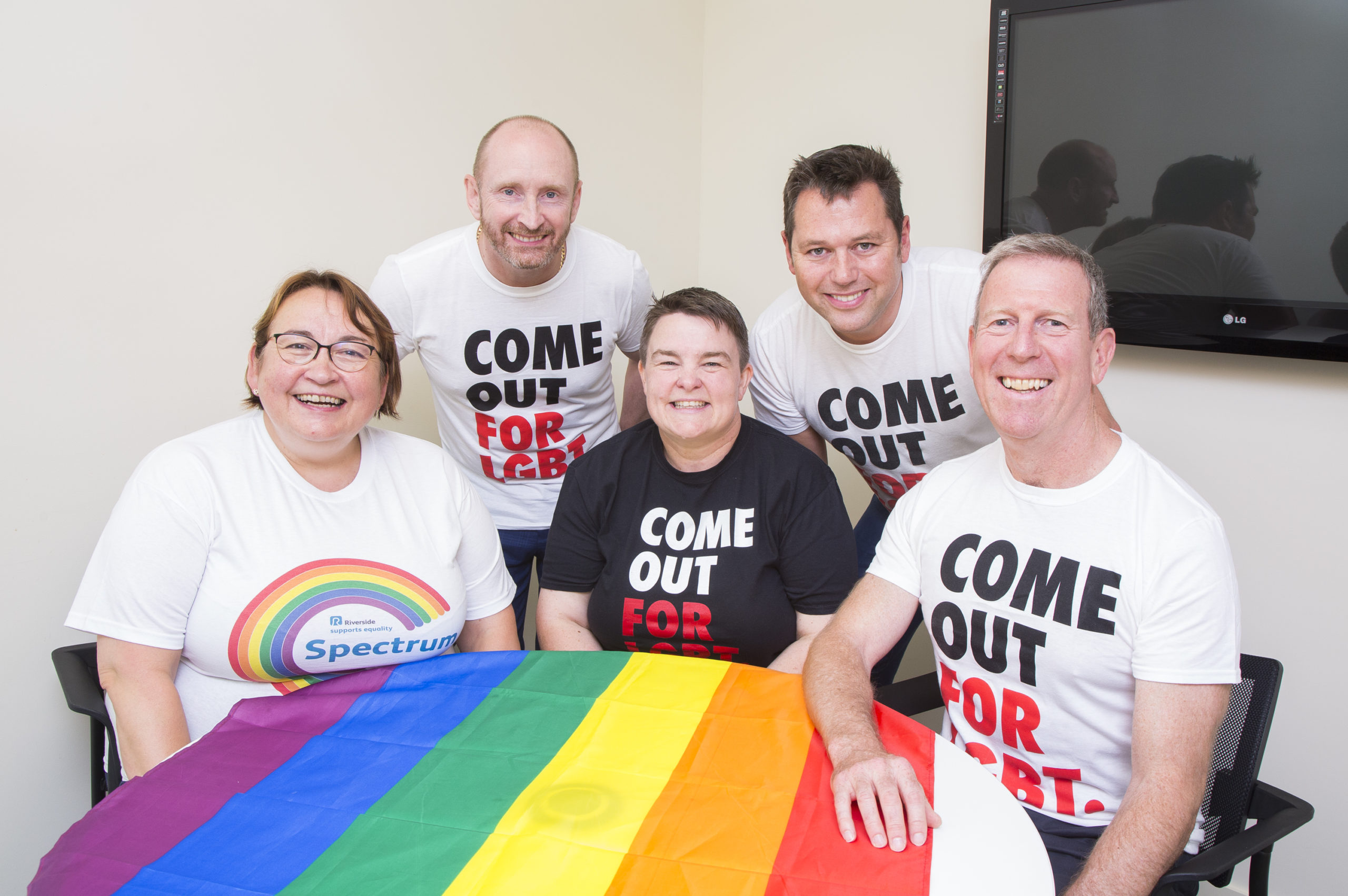 Group of five people seated and standing around a table covered with a rainbow Pride flag. They are wearing white T‑shirts with slogans including “Come Out For LGBT” and “Spectrum.” The group is indoors in a meeting room, with a screen mounted on the wall behind them.