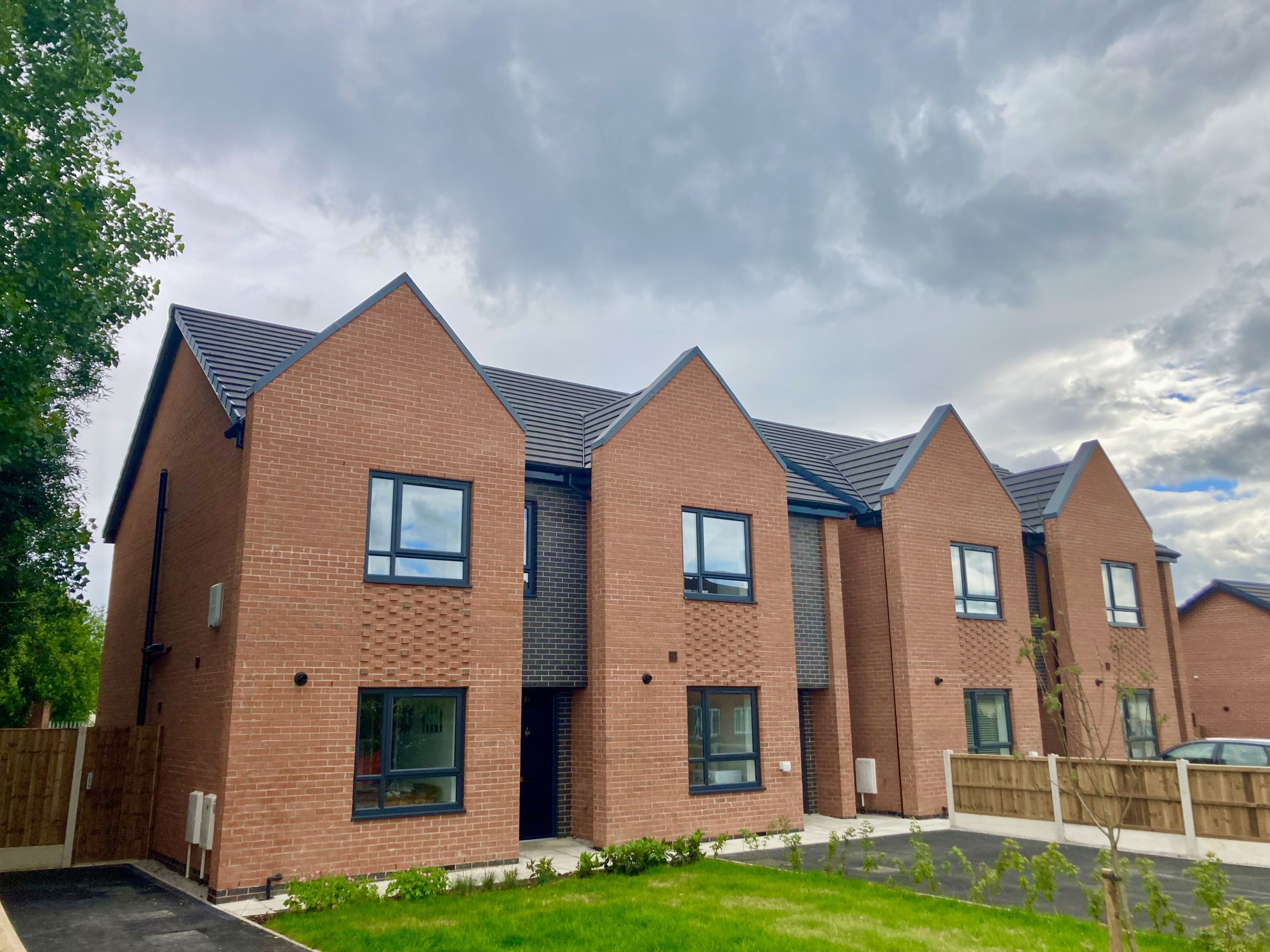 Row of newly built red‑brick houses with pitched roofs and dark window frames. A small grass lawn and paved path run along the front of the homes, with a cloudy sky overhead.