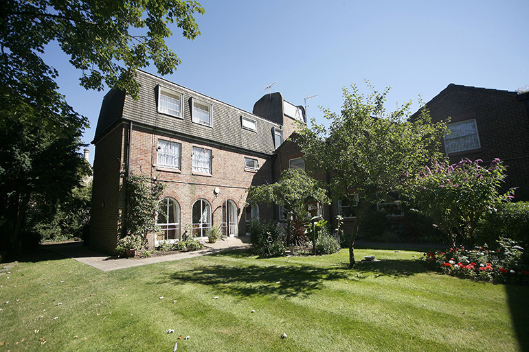 Exterior of a three‑storey brick residential building with arched ground‑floor doorways, viewed across a well‑maintained lawn and garden. Trees, shrubs and flower beds surround the building, with sunlight casting shade across the grass and façades.