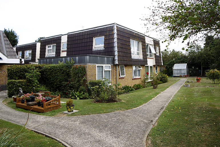 Exterior of a two‑storey residential building with brown cladding on the upper level and light brick walls below. A curving paved footpath runs through a landscaped communal garden with lawns, shrubs and flower beds, leading past seating areas and planters, with trees and greenery around the shared outdoor space under an overcast sky.
