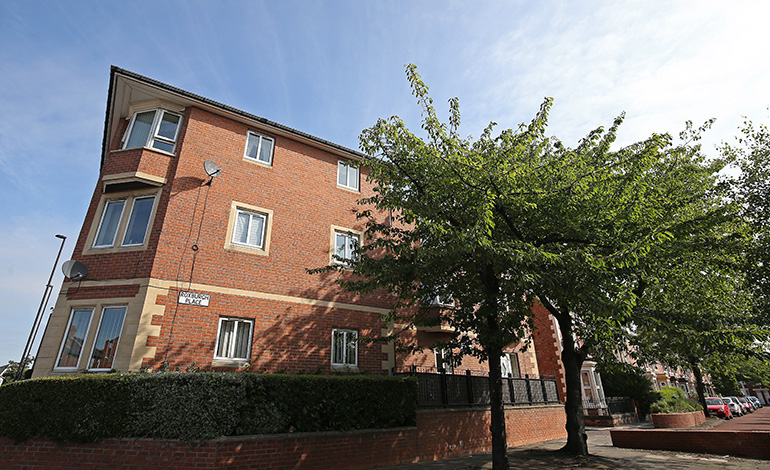 Exterior of a three‑storey red‑brick residential building with white‑framed windows, set along a quiet street. A mature leafy tree stands in the foreground beside a low brick wall and hedge, with the building façade and neighbouring homes visible under a bright blue sky.