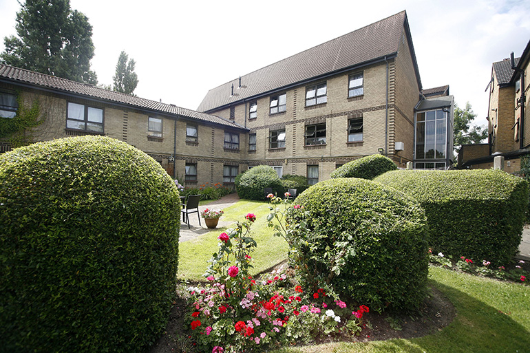 Communal garden courtyard surrounded by a multi‑storey light‑brick residential building with brown‑framed windows. Neatly trimmed hedges and rounded shrubs frame a central lawn with colourful flower beds and potted plants, while paved paths and seating areas create a calm shared outdoor space beneath a bright, lightly clouded sky.