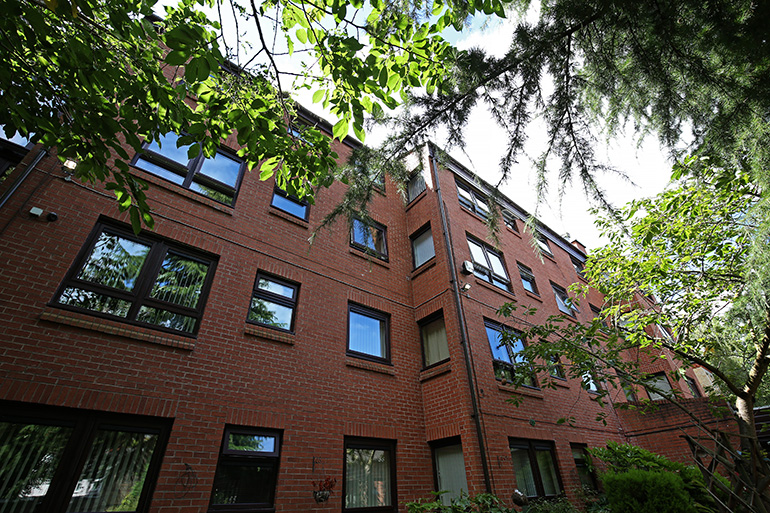 Exterior view looking up at a multi‑storey red‑brick residential building with wooden‑framed windows, partially framed by overhanging tree branches and leaves. The building is surrounded by mature greenery, with sunlight filtering through the trees against a partly bright sky.