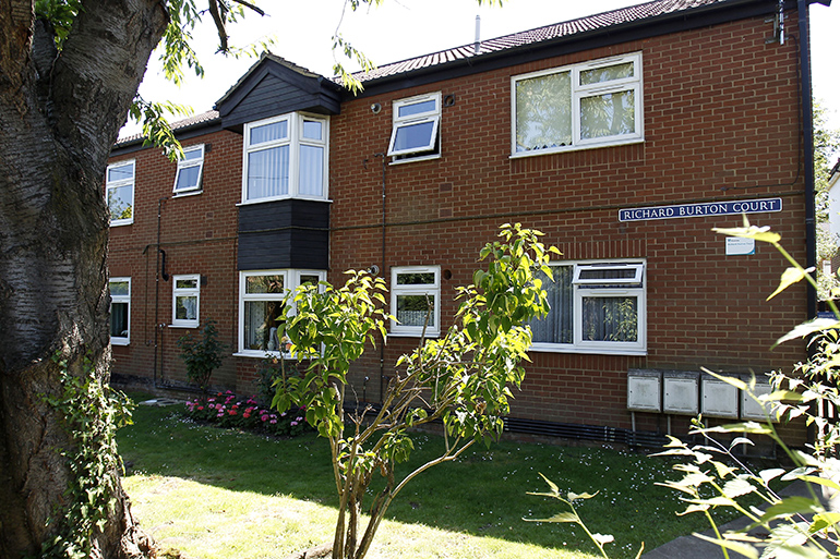 Exterior of a two‑storey red‑brick residential building with white‑framed windows, viewed across a small lawn with trees and shrubs. A blue street‑style sign on the building reads “Richard Burton Court,” with planted flower beds along the frontage and leafy branches framing the scene under bright daylight.