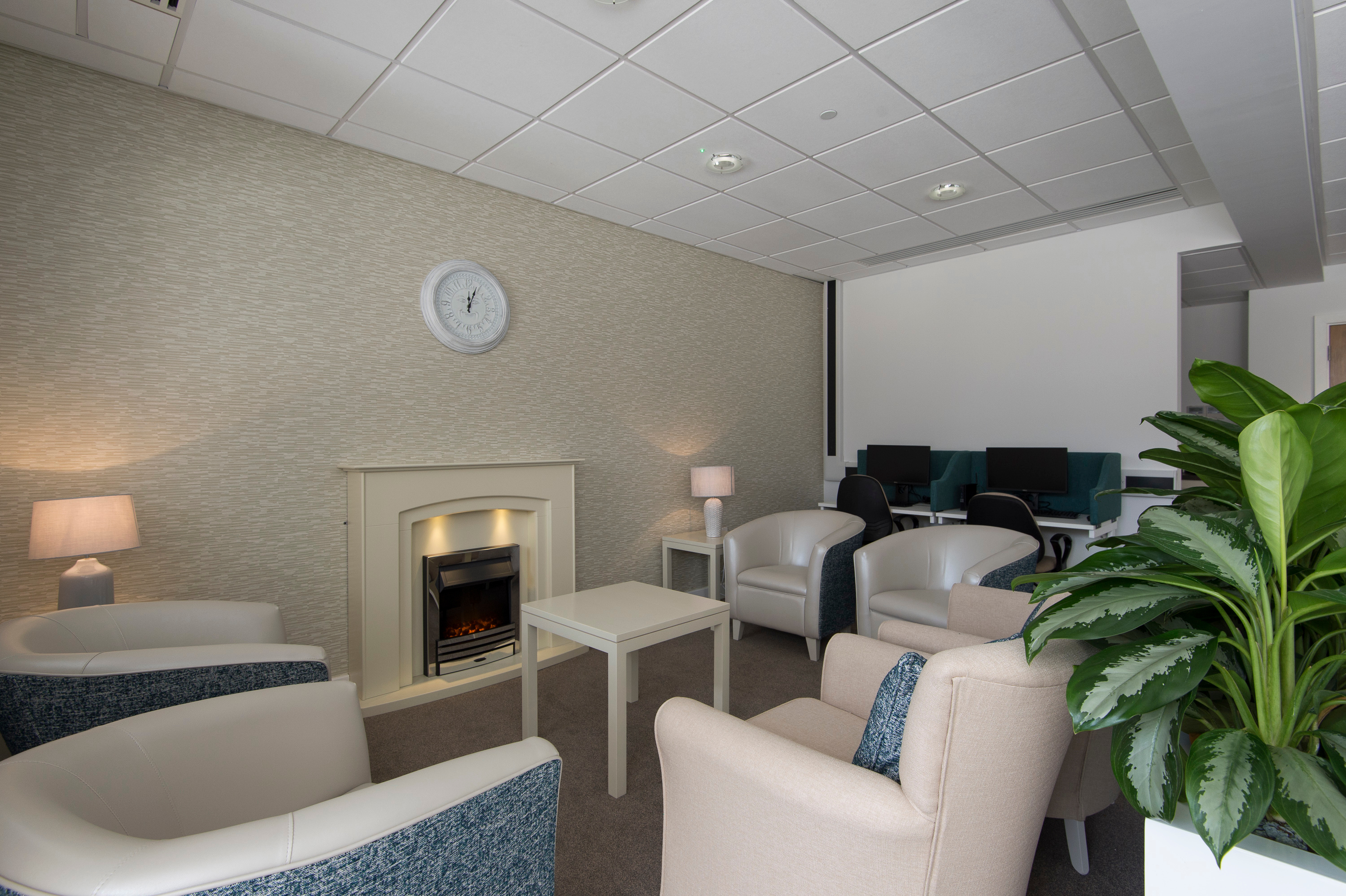 Quiet sitting area with armchairs around a coffee table, electric fireplace and desk workstations in the background.