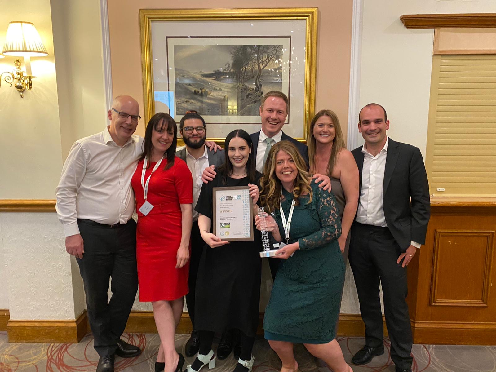 Group of people standing together indoors at an awards event, holding a certificate and a trophy. They are posing in front of a framed picture on the wall, with others gathered closely around them in a formal setting.