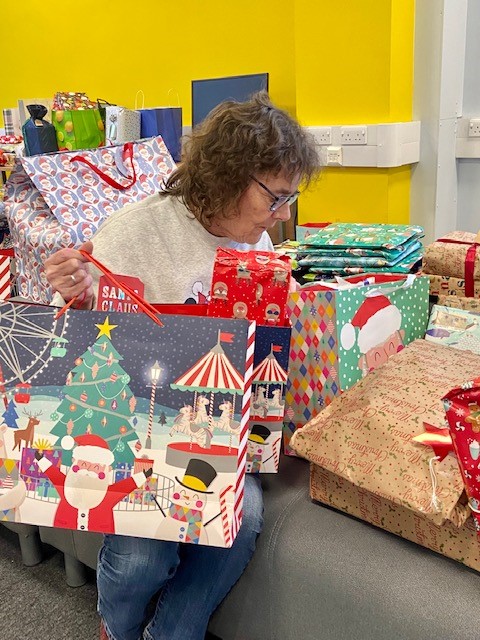 Engage Leeds Toy Appeal Christmas 2022 Jean Person seated indoors sorting Christmas gift bags and wrapped presents. Colourful festive gift bags, boxes and wrapping paper are arranged on a bench and table around them, with a bright yellow wall in the background.