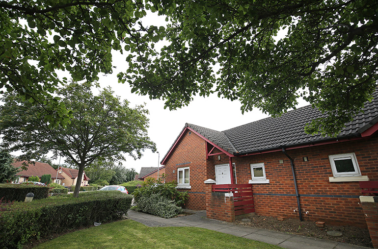 Exterior of a single‑storey red‑brick bungalow with a grey tiled roof, set within a quiet residential area. A paved path leads to the front door, with neatly trimmed hedges, small shrubs and a lawn in the foreground, tree branches overhead, and neighbouring bungalows visible in the background.