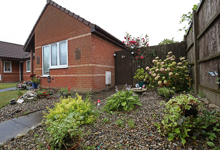 Small red‑brick bungalow with white‑framed windows, viewed from a gravel garden filled with plants and decorative features. Flowering shrubs, potted plants and a small water feature sit in the foreground, with a wooden fence and garden ornaments along the boundary and neighbouring homes visible behind.