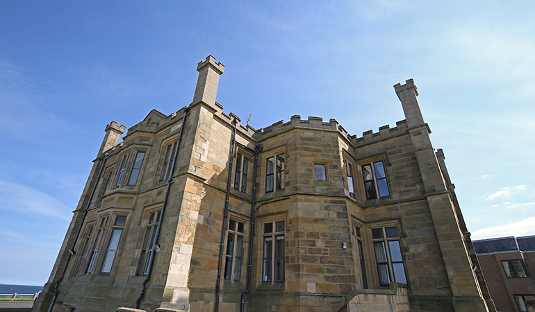 Exterior of a large historic stone building with multiple storeys, tall windows and castellated architectural details. The façade features projecting bays and chimneys, with light‑coloured stonework set against a clear blue sky.