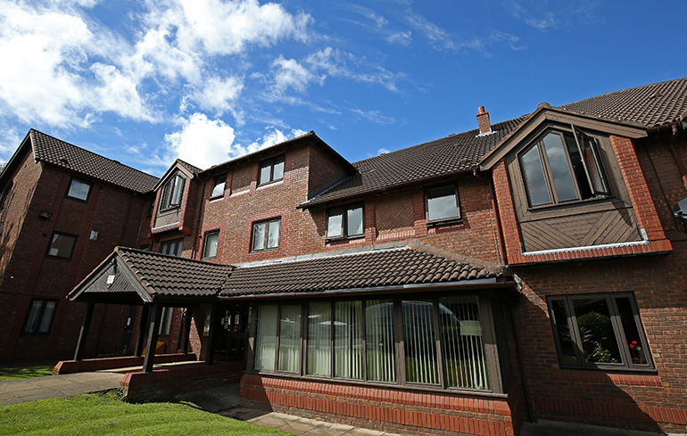 Exterior of a multi‑storey red‑brick residential building with pitched tiled roofs and white‑framed windows. A glazed single‑storey extension with vertical blinds faces a small lawn in the foreground, with a covered entrance canopy to one side and additional upper‑floor windows visible against a bright blue sky with scattered clouds.
