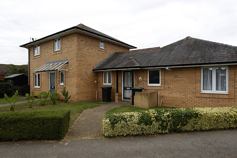 Exterior of a small group of low‑rise brick cottages with white‑framed windows and pitched roofs. A paved path curves past neatly trimmed hedges and a small lawn toward the entrances, with a taller central building behind the single‑storey homes under an overcast sky.