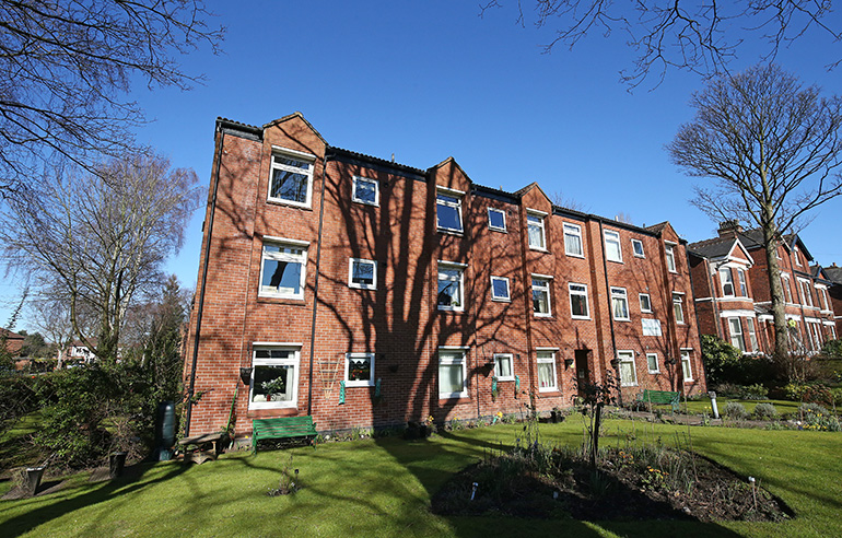 Exterior of a multi‑storey red‑brick residential building with rows of white‑framed windows, viewed across a neatly maintained garden. A large tree casts shadows across the lawn in the foreground, with flower beds, benches and mature trees surrounding the building under a clear blue sky.