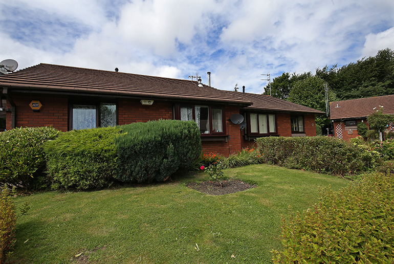 Exterior of a single‑storey red‑brick bungalow with white‑framed windows and a pitched tiled roof, set within landscaped gardens. A neatly kept lawn and trimmed hedges sit in the foreground, with low shrubs, flowering plants and a paved path around the building, and similar residential homes visible nearby under a partly cloudy sky.