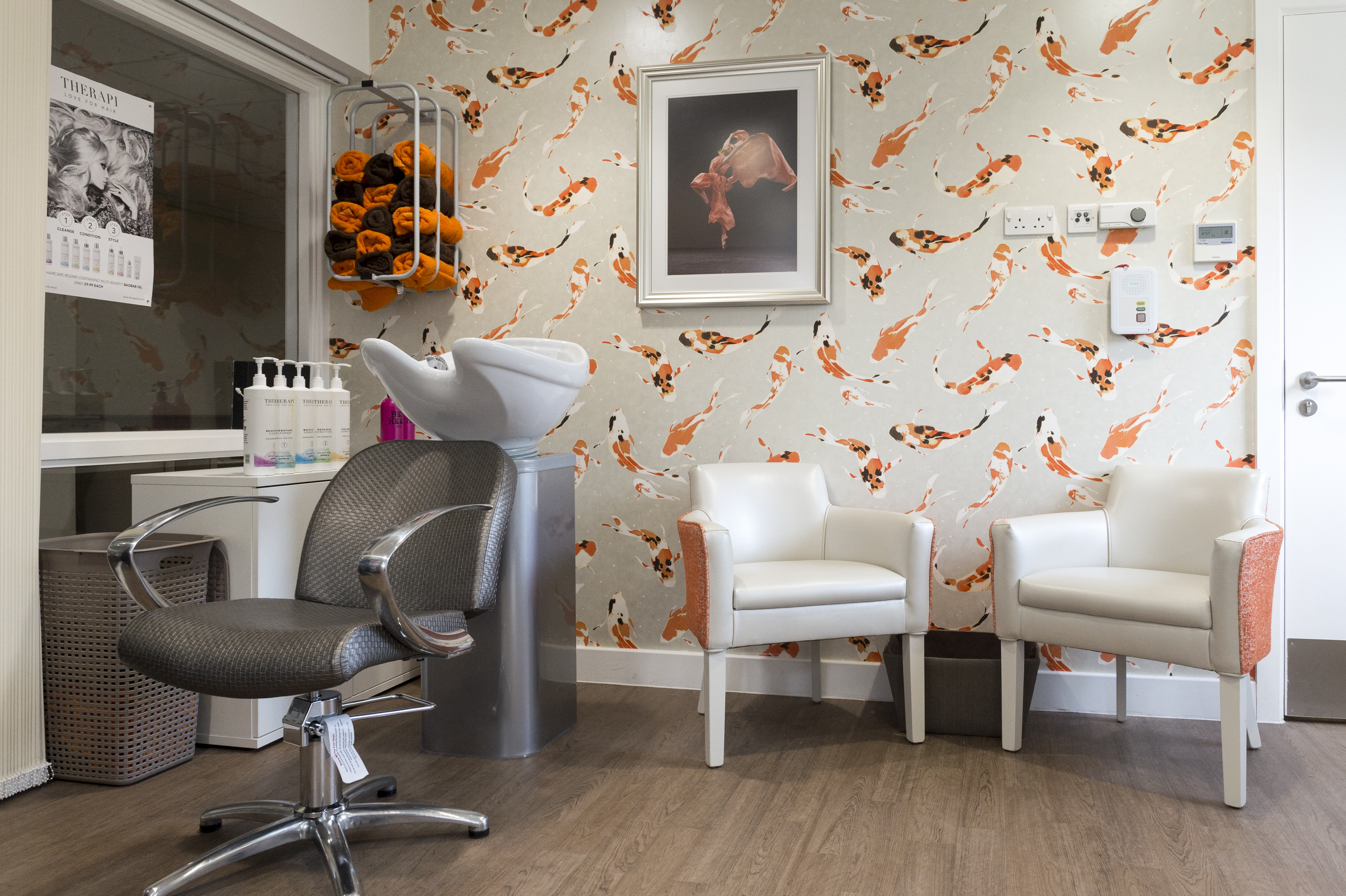 Interior of a salon-style room with a hair-washing sink, styling chair, shelves of hair products, folded towels, and patterned wallpaper, with two armchairs beside the sink.