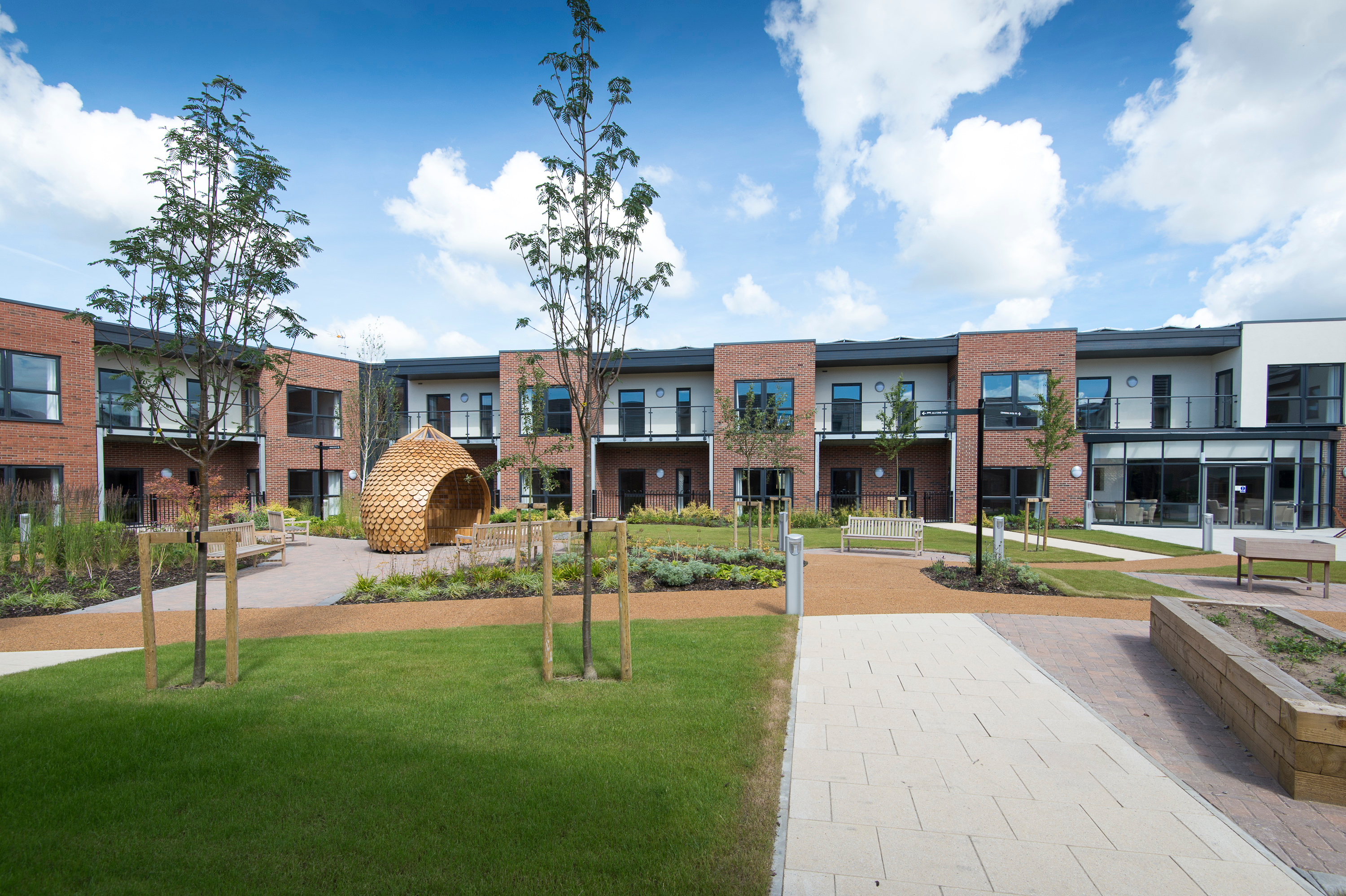Modern retirement housing courtyard with landscaped paths, benches, young trees and a wooden seating pod, surrounded by two‑storey brick apartments with balconies.