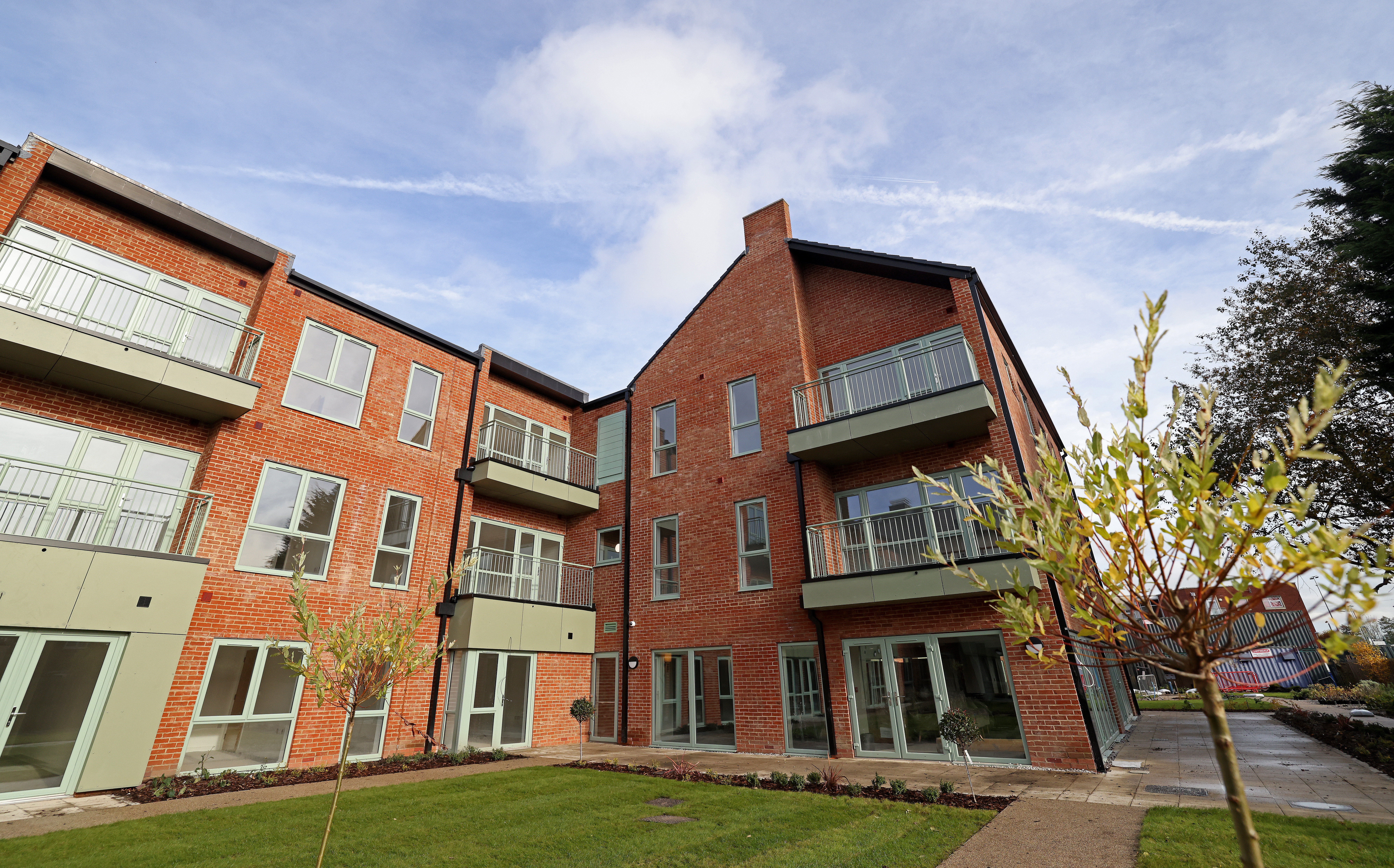 Red-brick apartment block with balconies, viewed from a landscaped courtyard with grass, paths and small trees.