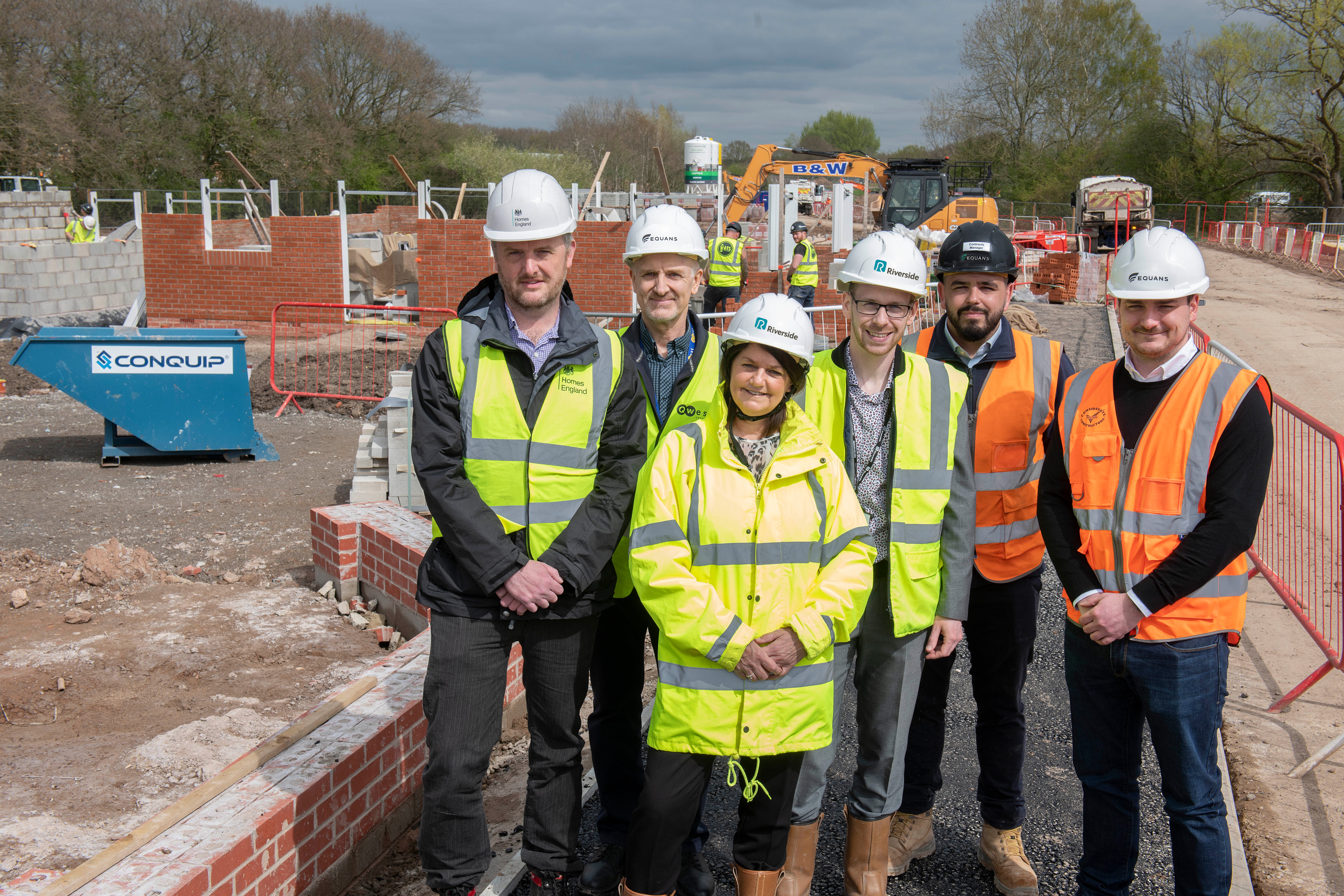 Group of people wearing high‑visibility jackets and safety helmets standing together at a construction site. A partially built brick structure, construction equipment and site fencing are visible behind them, with trees and cloudy sky in the background.