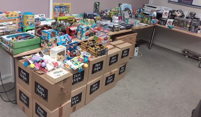 Tables filled with stacked cardboard boxes labelled “Box of Hope,” topped with a wide range of donated toys, games and gifts. The boxes and items are arranged neatly in an indoor room with carpeted flooring and noticeboards visible on the walls.