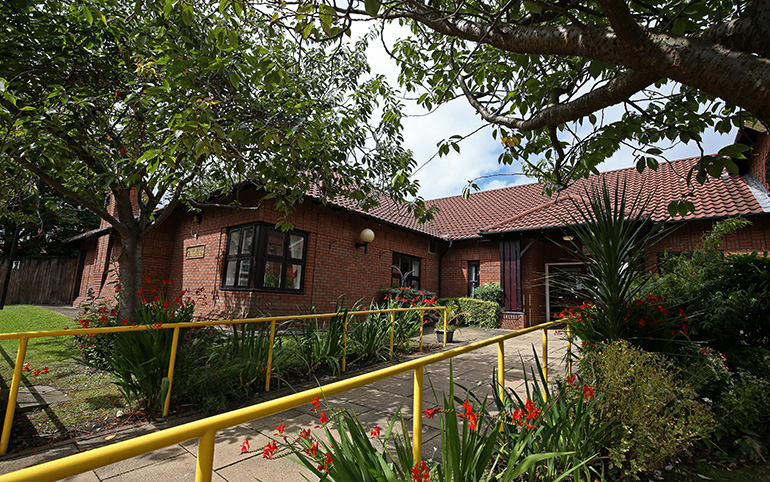 Single‑storey red‑brick residential building with a pitched tiled roof, surrounded by landscaped gardens. A paved pathway with yellow handrails leads to the entrance, with trees, shrubs and flowering plants around the building.
