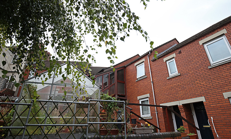 Exterior view of red‑brick terraced houses with white‑framed windows and front doors. A raised garden area with trellis fencing, potted plants and greenery sits in the foreground, with tree branches overhead and neighbouring buildings visible behind.