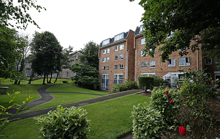 Large multi‑storey red‑brick residential building set within landscaped grounds. A winding paved path runs through lawns and planted shrubs, with mature trees and flowering bushes surrounding the building and windows overlooking the shared green space.