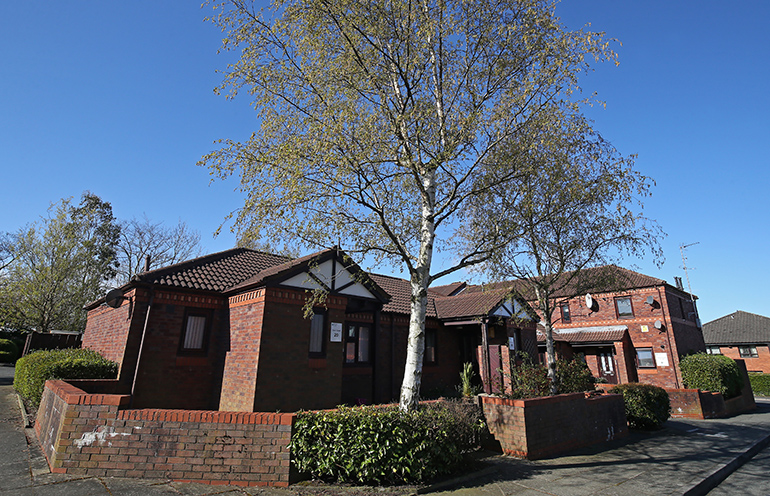Single‑storey red‑brick residential buildings set along a quiet street, with pitched tiled roofs and small entrance porches. Mature trees line the pavement and garden walls border the properties, with low shrubs and planting around the buildings under a clear blue sky.