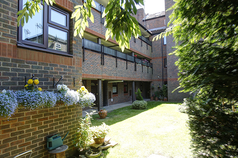Landscaped communal garden beside a red‑brick residential building, with a sunlit lawn, potted plants and low shrubs. Upper‑floor balconies overlook the garden, with trees and foliage framing the space and dappled sunlight across the grass and brickwork.