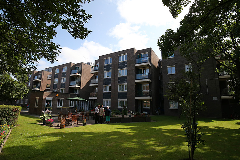 Landscaped communal garden in front of a modern multi‑storey residential building with balconies. A large lawn stretches across the foreground, with trees framing the space and raised planters near seating where a small group of residents are gathered outdoors under a bright sky.