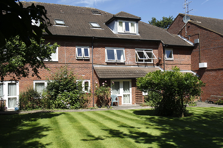 Exterior of a two‑storey red‑brick residential building with white‑framed windows and a central entrance beneath a small tiled canopy. The building faces a neatly mown lawn with shrubs and plants along the frontage, bordered by trees and greenery under a bright blue sky.