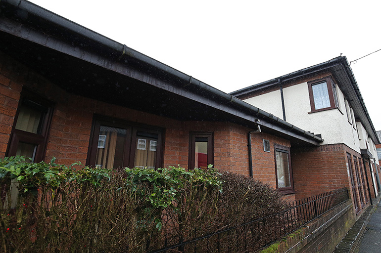 Exterior of a two‑storey residential building with red brick on the ground floor and white‑rendered upper walls. White‑framed windows line the building, with a narrow pavement running alongside a low hedge and metal railing, and neighbouring buildings visible along the street under an overcast sky.