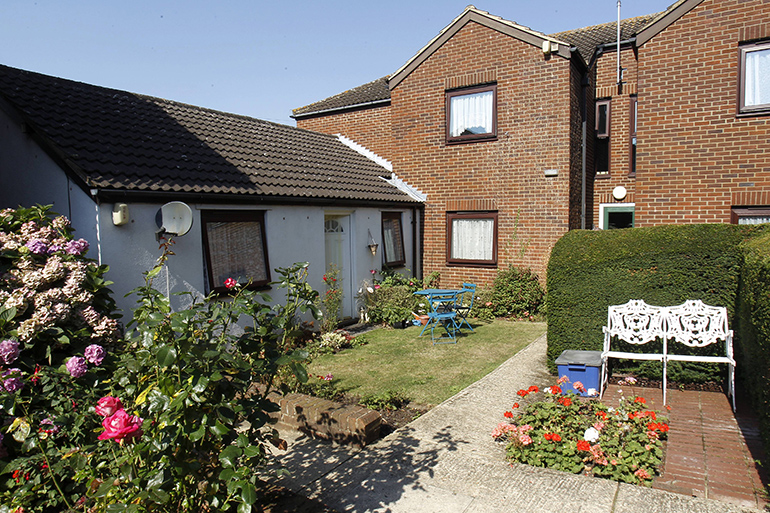 Communal garden courtyard between red‑brick residential buildings, featuring a paved path, trimmed hedges, flower beds and outdoor seating. A small lawn area with plants and a blue table and chairs sits outside ground‑floor homes, creating a well‑maintained shared space.