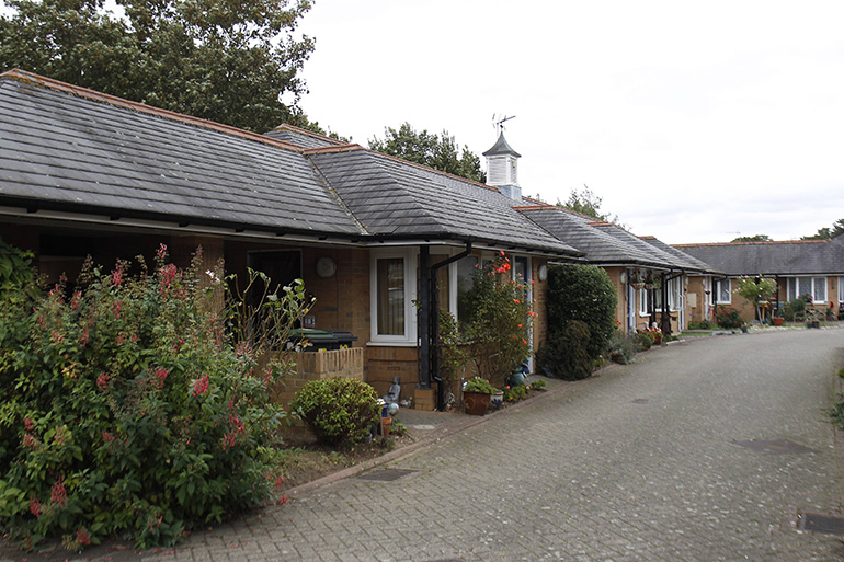 Row of single‑storey brick cottages with pitched tiled roofs, arranged along a quiet paved access road. Each home has a small covered porch and front garden with shrubs and potted plants, creating a calm residential setting with trees in the background.
