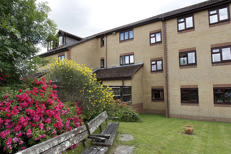 Communal garden beside a multi‑storey light‑brick residential building with white‑framed windows. A wooden bench sits on a neatly kept lawn in the foreground, bordered by flowering shrubs and bushes in pink and yellow bloom, with trees and planting creating a calm shared outdoor space.