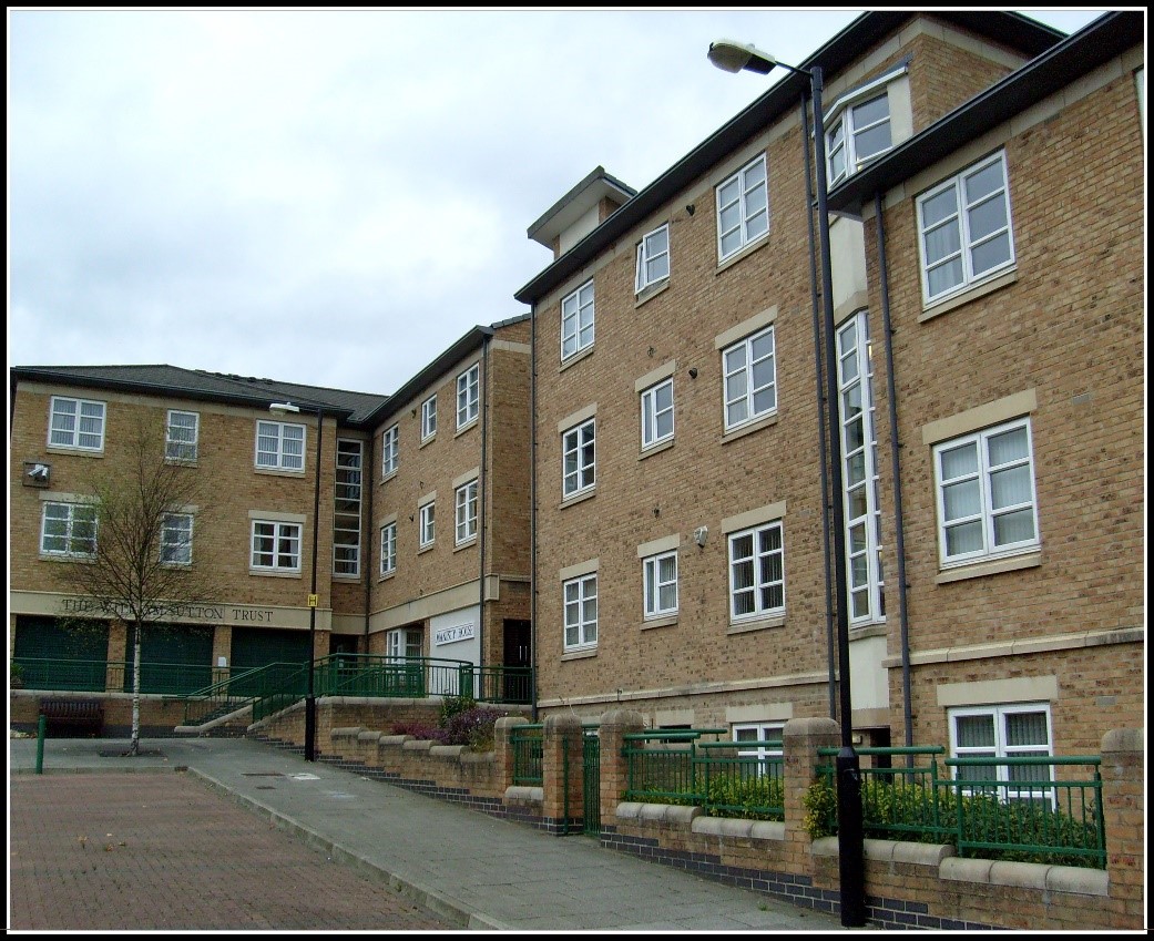 Exterior of a multi‑storey light‑brick residential building with white‑framed windows, arranged around a paved communal area. A ramp with green metal railings leads to the entrance, with low brick walls, planted borders and a streetlamp visible in the foreground, under an overcast sky.