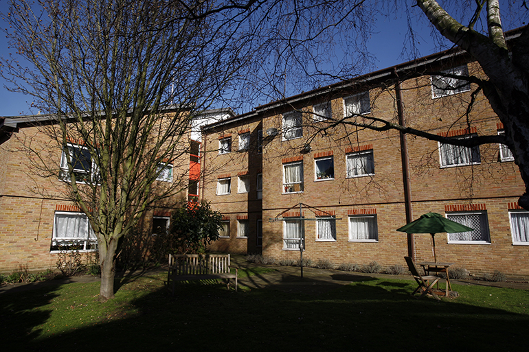 Communal garden courtyard in front of a multi‑storey light‑brick residential building with white‑framed windows. A neatly kept lawn sits beneath leafless trees casting shadows across the grass, with wooden benches and a small table with a parasol placed in the garden. Ground‑floor windows look onto the shared outdoor space, with shrubs and planting around the edges under bright sunlight and a clear blue sky.
