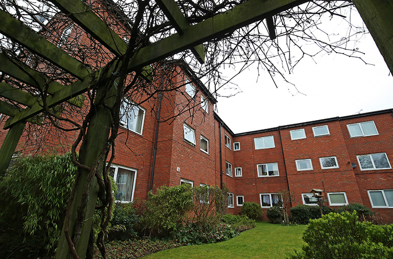 Communal garden courtyard beside a multi‑storey red‑brick residential building with white‑framed windows. A wooden pergola with climbing plants frames the foreground, with a neatly kept lawn, shrubs and flower beds below, and the building surrounding the shared outdoor space under an overcast sky.