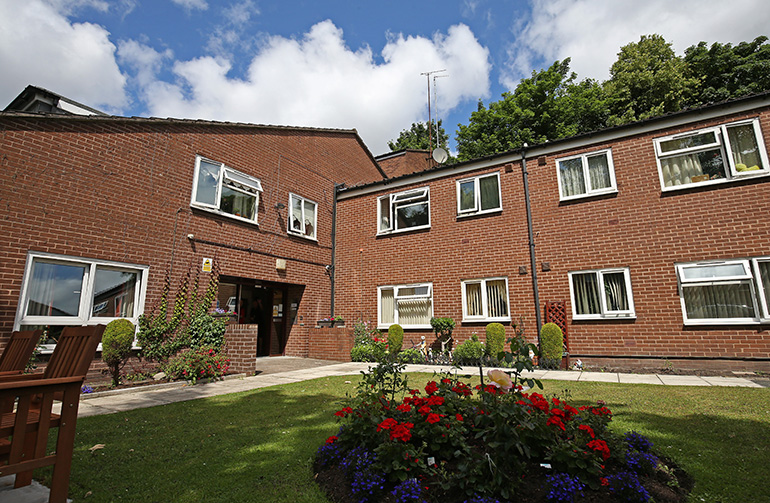 Exterior of a two‑storey red‑brick residential building arranged around a landscaped communal garden. A circular flower bed with red and blue flowers sits in the foreground on a neatly kept lawn, with paved paths, benches and white‑framed windows overlooking the shared outdoor space under a partly cloudy sky.