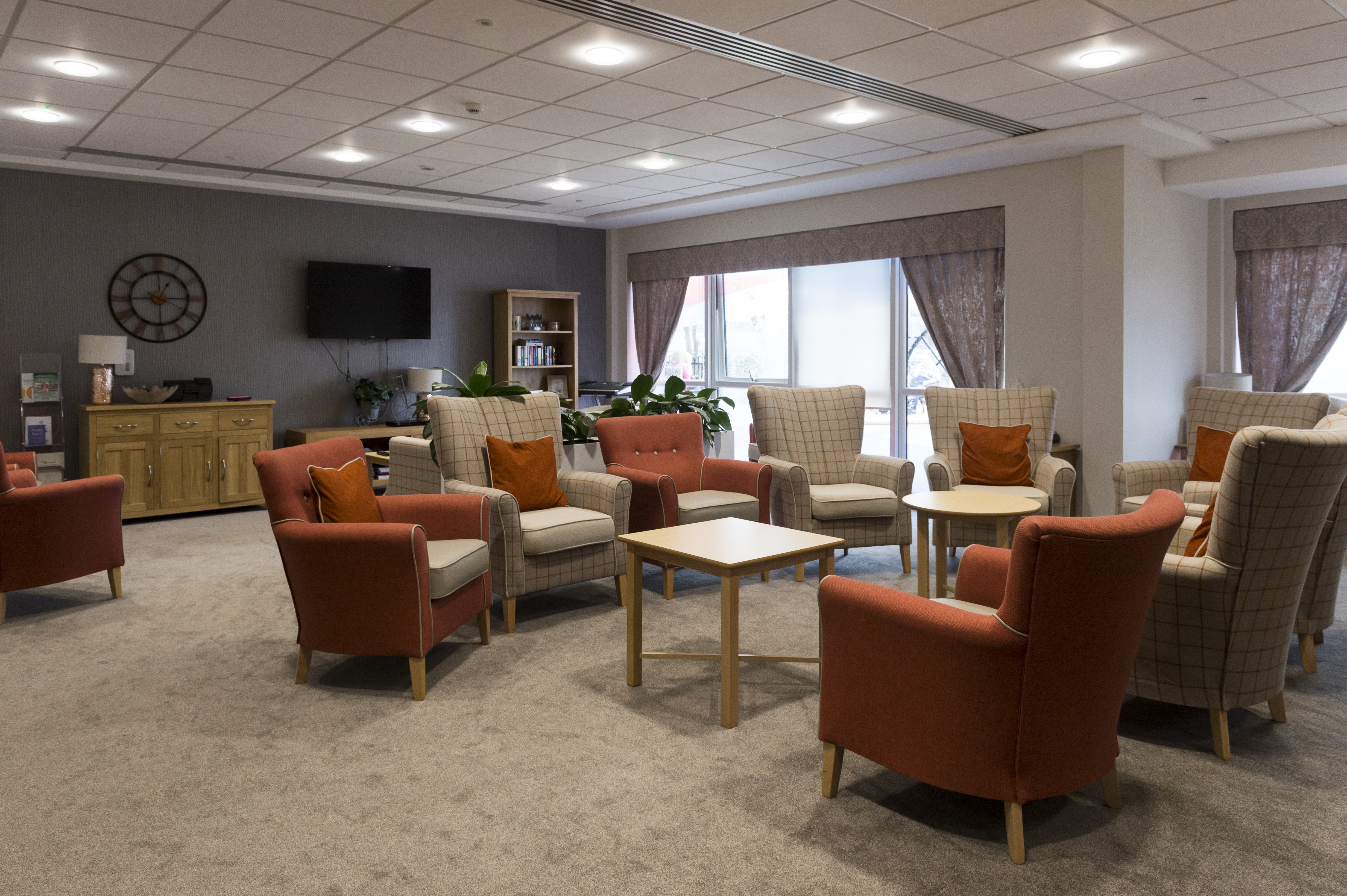 Lounge area with multiple armchairs arranged in a circle around small tables, with a television, shelving, and large windows letting in natural light.