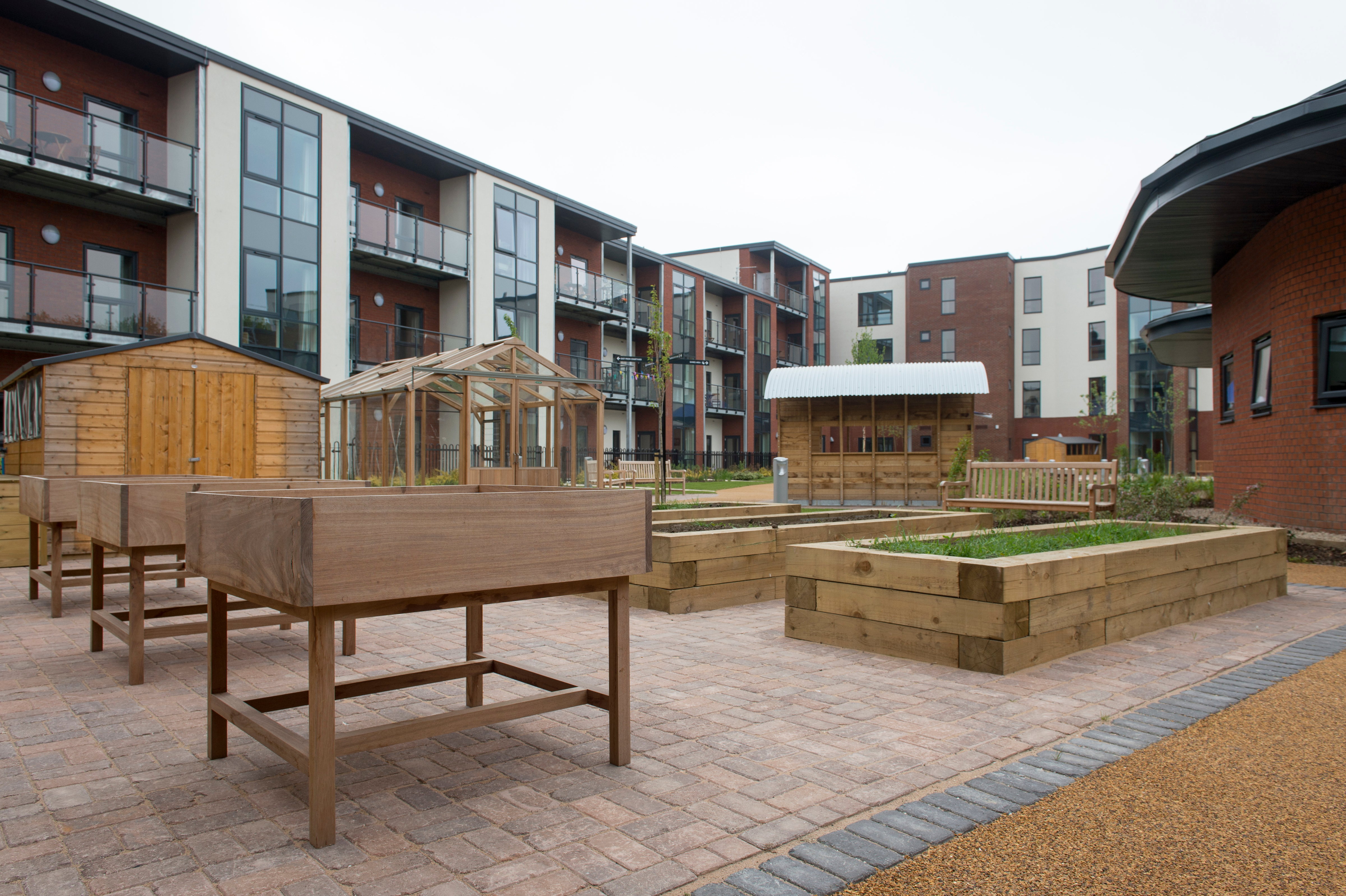 Accessible courtyard garden with raised planting beds, benches, a greenhouse and wooden sheds, surrounded by modern apartment buildings with balconies.