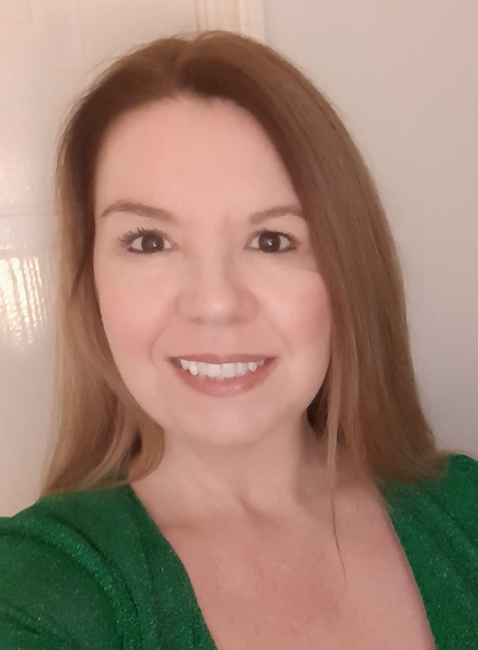 Portrait of a person with long, light brown hair facing the camera, wearing a green top. The photo is taken indoors against a plain background with a door visible behind them.