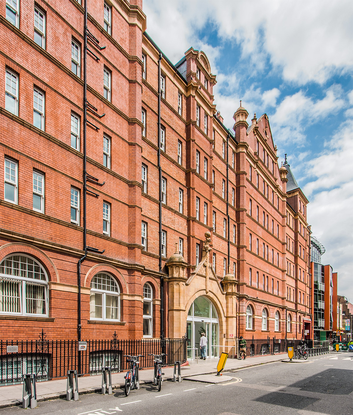 Exterior view of a large red‑brick historic building with multiple storeys and arched windows. A central arched entrance faces the street, with bicycles parked along railings outside and a partly cloudy sky overhead.