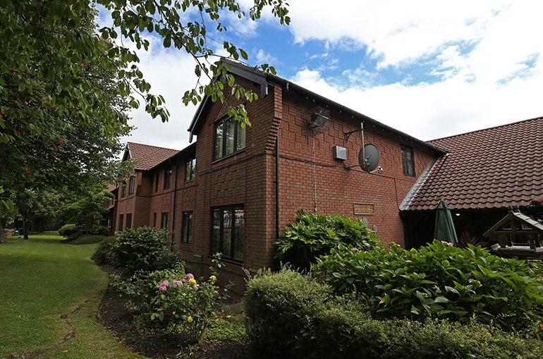 Two‑storey red‑brick residential building set within a landscaped garden. Shrubs, flowering plants and lawn areas surround the building, with trees framing the scene under a partly cloudy blue sky.