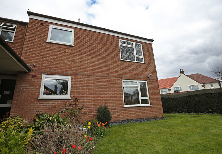 Exterior of a two‑storey red‑brick residential building with white‑framed windows, set beside a neatly maintained lawn. Flowering plants and shrubs line the base of the building, with neighbouring houses visible beyond hedging under a cloudy sky.