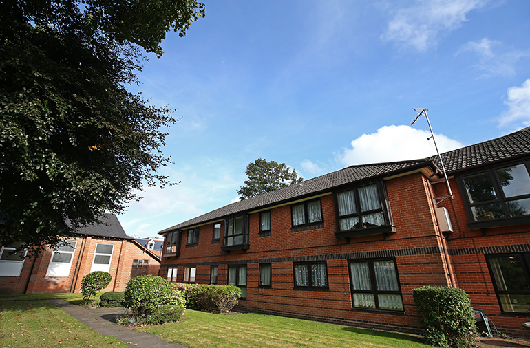 Exterior of a two‑storey red‑brick residential building with white‑framed windows, set beside a neatly maintained lawn. Low shrubs line a paved footpath leading past the building, with mature trees providing shade and another residential block visible in the background under a blue sky.
