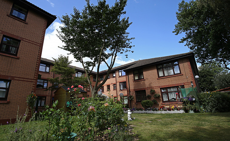 Landscaped communal garden in front of a two‑storey red‑brick residential building with white‑framed windows. Flower beds with roses and other plants fill the foreground, with a neatly kept lawn, small garden ornaments and shrubs, and mature trees providing shade around the shared outdoor space under a blue sky.