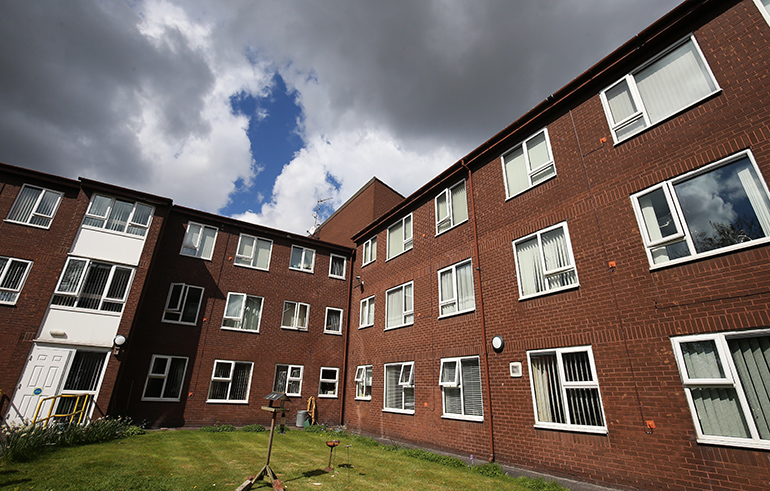 Exterior of a large multi‑storey red‑brick residential building with white‑framed windows, arranged around a communal lawn. Bird tables and small garden features are positioned on the grass, with paths and planted borders around the courtyard, and dramatic dark clouds breaking to blue sky overhead.
