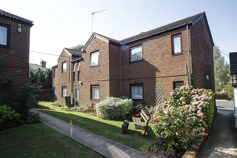 Exterior of a two‑storey red‑brick residential building set around a landscaped communal garden. A paved footpath runs through a neatly kept lawn with shrubs, flowering plants and a wooden bench, with windows facing the shared outdoor space under clear daylight.